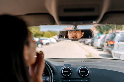 Woman applying lipstick in car, reflected in a rear view mirror