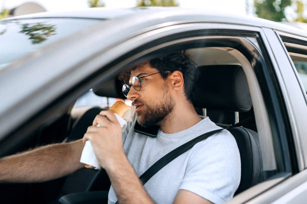 istock Young casually clothed man talking on a phone and eating sandwich while driving car 1633255417