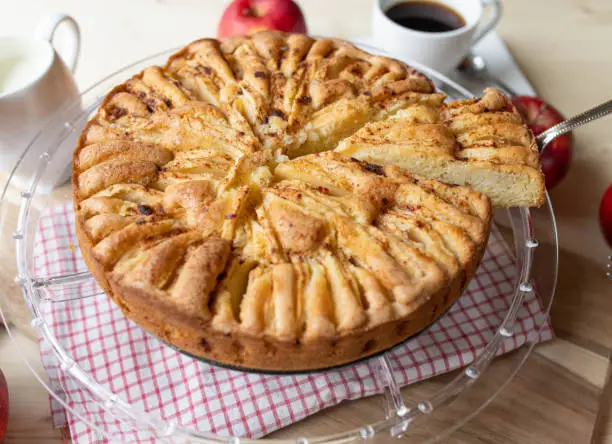Homemade apple pie with cinnamon topping. Served whole with a slice on a cake server on rustic table with coffee and fresh apples.