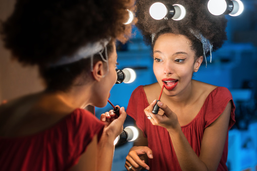 Young Moroccan female with Afro hair headband looking away while standing in front of makeup mirror with illuminated lights and putting red lipstick against blurred background