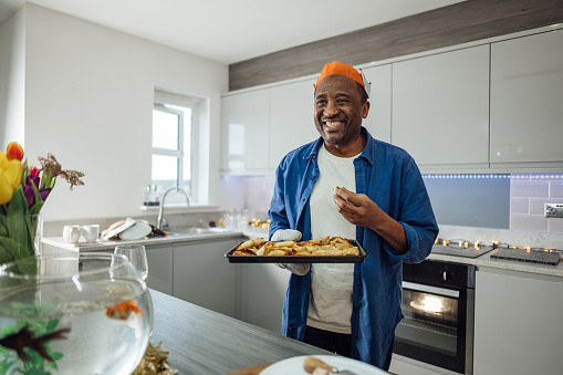 A senior man standing in the kitchen in his house on Christmas Day in Gateshead, North East England. He is wearing a paper crown and holding a tray of freshly cooked samosas while looking away from the camera and smiling.