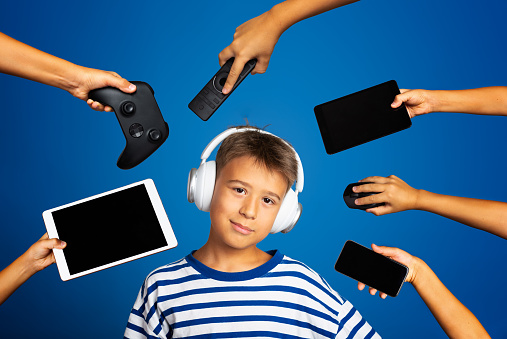 Teenage boy with headphones , surrounded by hands offering him  phones, joystick, remote control, tablet...