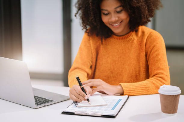 istock Business woman using laptop to work and writing on notebook with pen in office with mobile next to laptop. 1633429955