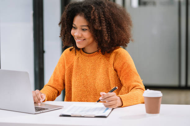 istock Business woman using laptop to work and writing on notebook with pen in office with mobile next to laptop. 1633430267