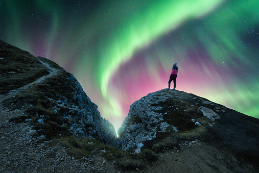 Northern lights and young woman on mountain peak at night. Green and violet aurora borealis and silhouette of alone girl on mountain trail. Landscape with polar lights. Starry sky with bright aurora