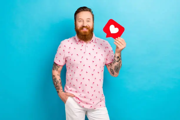 Photo of Photo of satisfied positive man with redhair beard wear pink t-shirt hold social media like icon isolated on blue color background