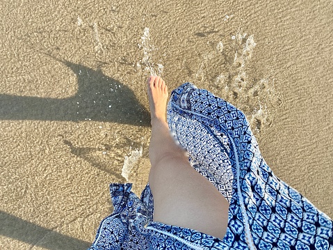 Horizontal seascape looking down to barefoot woman walking sand shoreline in long blue patterned dress split dress on windy day at Byron Bay Australia