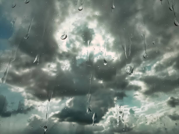 A close-up photo of rain drops falling on a window. Dark and stormy skies in the background. The rain drops are large and heavy, and running down the window.