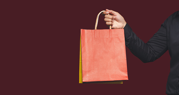 Close-up of hand holding a red paper bag with a handle while standing on a red background. Packaging template mockup. Delivery service, shopping, and advertising area concept