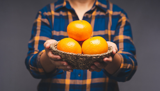 Close-up of hands holding a basket with fresh oranges while standing on a gray background. Healthy fruit concept