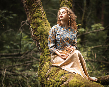 A young girl in shiny clothes sits on a moss-covered tree and looks dreamily to the side, the end of a happy youth. High quality photo