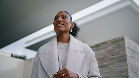 Closeup smiling black hair woman posing at modern house. Stylish girl wait taxi holding paper bags. Happy african american real estate agent looking camera at luxurious mansion. Successful businesswoman at home