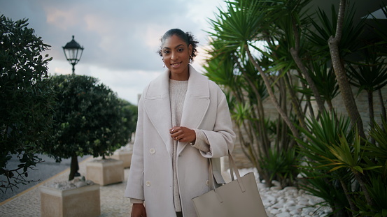 Attractive african american posing street closeup. Smiling woman hold shopping bags at contemporary luxurious residential area. Joyful black hair businesswoman in stylish coat. Wealthy elegant lady.