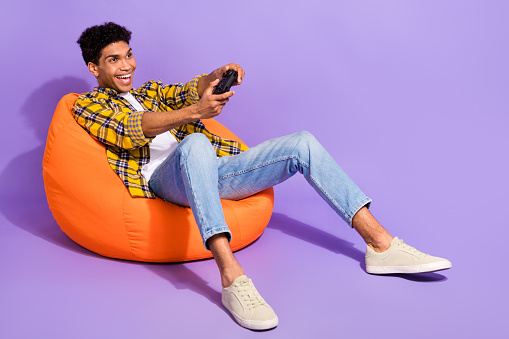 Full size portrait of overjoyed man sit comfort bag hold controller plating video games empty space isolated on purple color background.