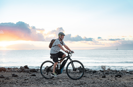 Happy active senior cyclist man at sea at sunset light with electric bicycle running on the beach - elderly man with helmet enjoying healthy lifestyle and freedom