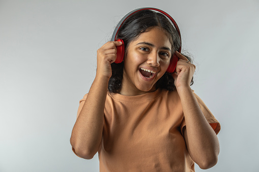 Portrait of cheerful teenage girl screaming and listening music over headphones while standing against white background