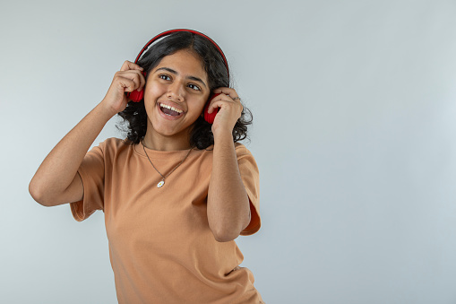 Portrait of cheerful teenage girl listening music over headphones while standing against white background