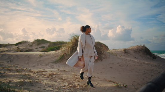 Single woman walk sand shore on spring vacation. Curly traveler admire ocean view lean wooden pier. Pensive relaxed african american stroll beach enjoying solitude. Calm mood weekend leisure concept