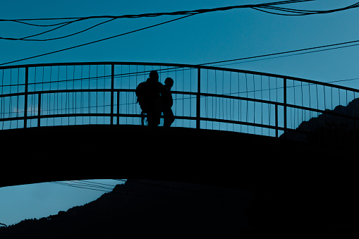 Silhouette of a man and his daughter watching the nature from a bridge above the road, at dawn, with a beautiful blue colored sky on the background, mountains and also electricity powerlines, specific for rural areas of Romania and Eastern Europe.