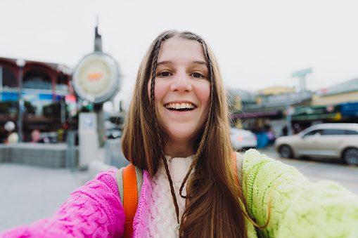 Portrait of smiling female with long hair and backpack wearing colourful knitted sweater exploring San Francisco city and taking photo of herself in old wharf by the sea