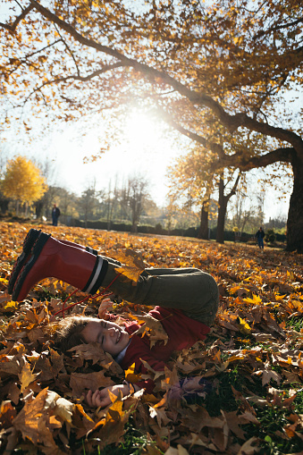 Side view of playful little girl with red rain boots lying in dry maple leafs in the park