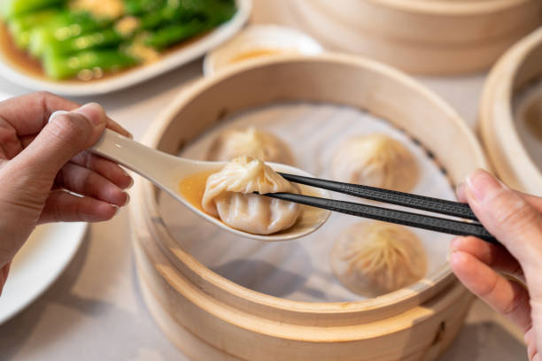 Person taking a dumpling from spoon with chopsticks. Closed up freshly steamed baozi dumplings in white spoon with bamboo steamer background.
