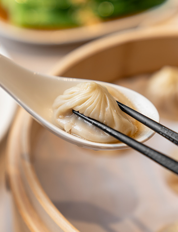 Person taking a dumpling from spoon with chopsticks. Closed up freshly steamed baozi dumplings in white spoon with bamboo steamer background.