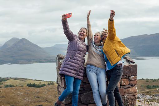 A medium close up of a group of three women who are proud of their achievement of getting to the summit of Applecross Peninsula in the Scottish highlands. They have made it to the summit and completed the challenge for themselves. They are holding up their arms with glee.