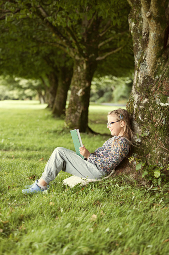 Fair-haired girl wearing spectacles reading her book under a tree.