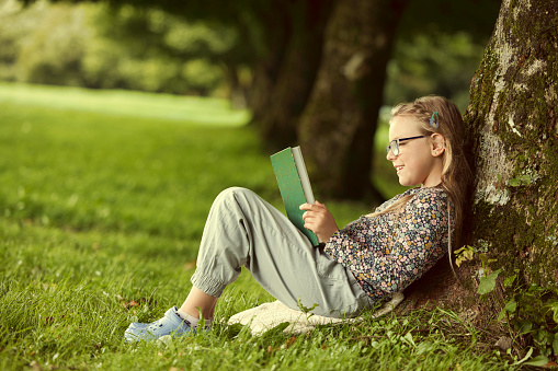 Beautiful smiling teenage girl in blue blouse lying on grass and read book, against green of summer park.