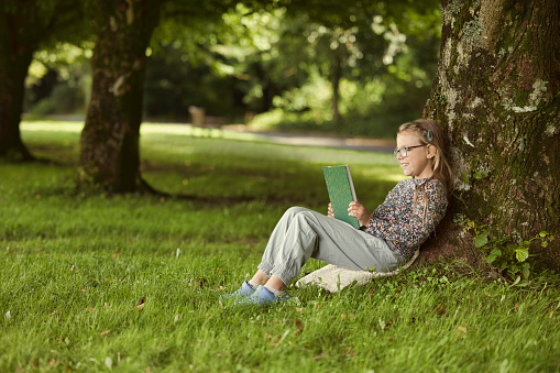Fair-haired girl wearing spectacles reading her book under a tree.