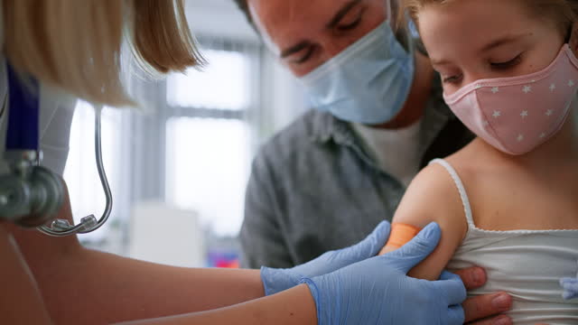 Little girl sitting on fahter's lap and getting vaccinated in doctor's office.