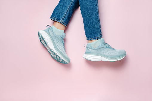 Women legs in blue jeans and new green blue modern running shoes on pink background. Stylish monochrome shoes for active people that incorporate new health technology. Top view
