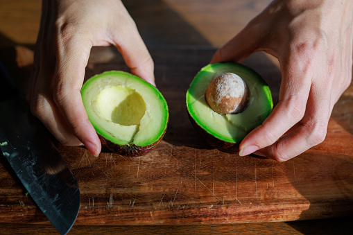 Close-up of female hands holding a ripe open avocado and preparing a healthy breakfast in the kitchen, with the beautiful morning sun shining on the avocado.