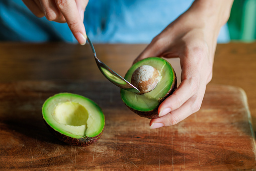 A close-up photograph capturing a woman skillfully peeling an avocado with a spoon on a cutting board, with the beautiful morning sun shining on the avocado.