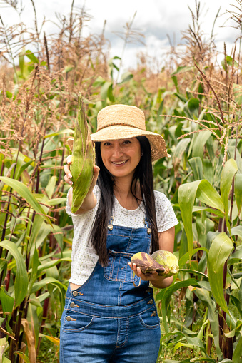 mujer joven vestida como jardinera indicando una mazorca de maiz que recien cosecho de su huerta