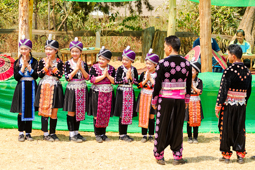 Rural thai community festival and traditional clothing scene in Chiang Mai province in small village and town area. People are wearing local traditional clothings based on black with colorful elements. Teenage girls are standing in a row and are looking into same direction.  They have folded hands for wai gesture,.They are preparing for local dance performance. Men in same fashion style are standing in front. In background village women are sitting and watching.