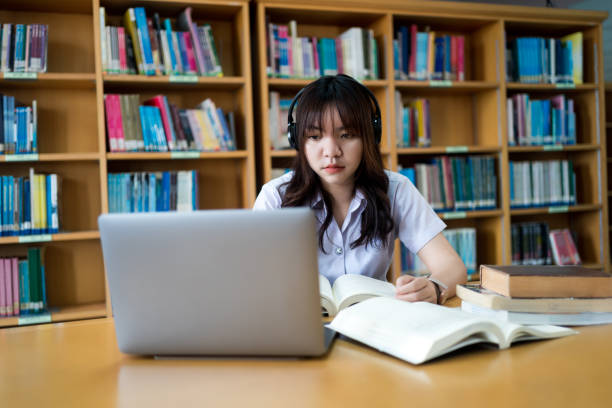 istock Young Asian female university student concentrate on studying, doing assuagement project in library with books and laptop on table 1633819168