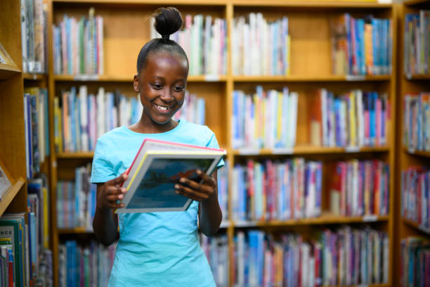 menina segurando um livro de histórias na biblioteca - africa child reading african descent - fotografias e filmes do acervo