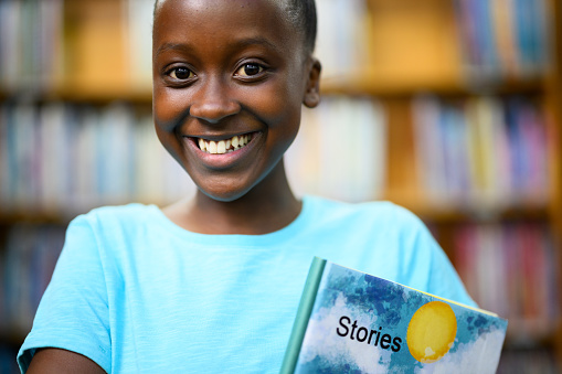 Happy young African girl holding a storybook in her arms at the library