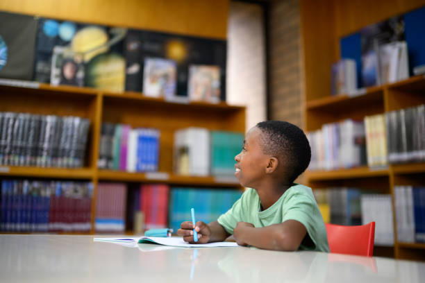 istock Side view young boy sitting at desk in  library looking up contemplating the answer 1633848349