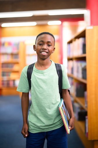 Young smiling African boy wearing his backpack standing in the library holding a book