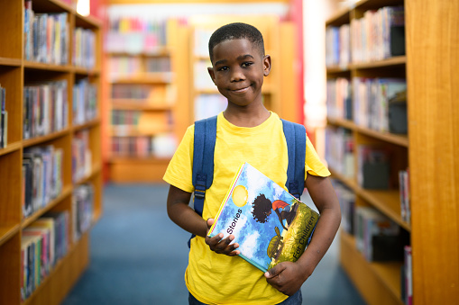 Young smiling African boy wearing his school backpack standing in the library between the bookshelves holding a book