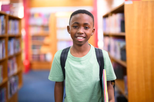 istock Portrait boy with rucksack holding a book standing in library between bookshelves 1633855890