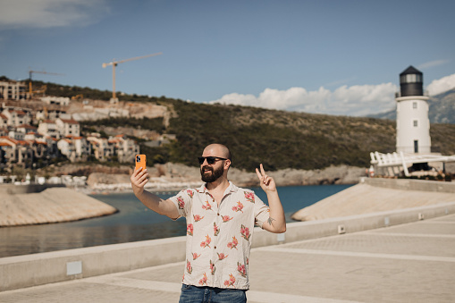 handsome bearded man wearing a Hawaiian shirt taking a selfie with a smartphone during his summer vacation at the seaport