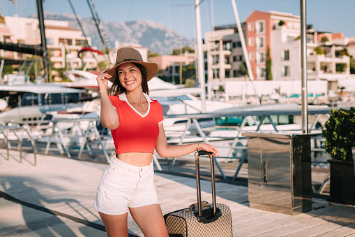 Portrait of a beautiful young casually dressed woman wearing a hat and carrying a suitcase at the seaport on a lovely sunny day during her summer vacation at the seaside