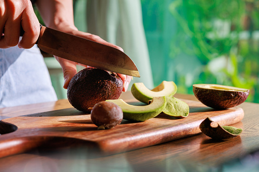 Close-up of female hands slicing a ripe avocado and preparing a healthy breakfast in the kitchen, with the beautiful morning sun shining on the avocado