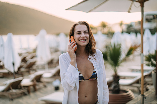 Portrait of a beautiful young woman wearing bikini and a white shirt using a smartphone at the beach during her summer vacation at the beach