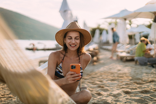 Beautiful young woman wearing a bikini using a smartphone and relaxing in a hammock chair at the beach and enjoying her summer vacation at the seaside
