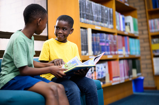 Two happy young African boys sitting in the library looking & talking about the book they are reading